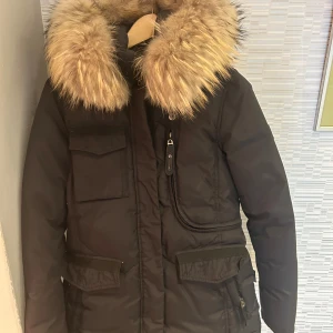 Svart dunjacka med päls från Hollies - Snygg svart dunjacka från Hollies med stor fluffig päls runt kragen och huva. Jackan har flera fickor framtill, dragkedja och snygga detaljer. 