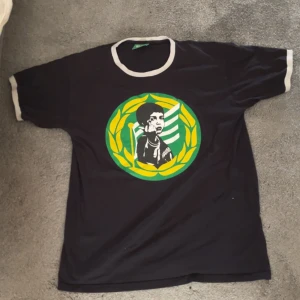 Svart Bajen Fans t-shirt med tryck - Svart t-shirt från Bajen Fans med vit kant vid hals och ärmslut. Stort tryck på bröstet med gul och grön krans samt motiv av en person. Skön passform och tillverkad i 100% bomull. Perfekt för dig som vill visa ditt stöd för Bajen.