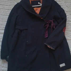 Odd Molly svart kappa med broderier - Svart kappa från Odd Molly med broderade röda rosor på ärmar och rygg, samt dekorativt vitt broderi på ryggen. Kappan har lila knytband i midjan och en orange blommig insida i sidenliknande material. Snygg, unik och med fina detaljer som spetskant och fickor.