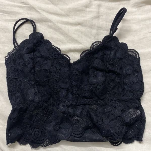 Svart spetsig croppad linnetopp - Supersöt svart croppad linnetopp från Lierrose med tunna axelband och blommig spets över hela toppen. Toppen har en feminin scallop-kant nertill och är perfekt för dig som gillar detaljer och romantisk stil.