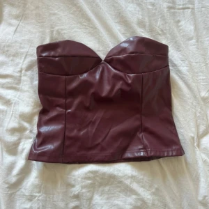 Vinröd skinntopp - Snygg vinröd tubtopp i skinnimitation med hjärtformad byst och slät finish. Toppen är axelbandslös och har en mjuk insida för extra komfort. Perfekt för dig som vill sticka ut med en edgy look.