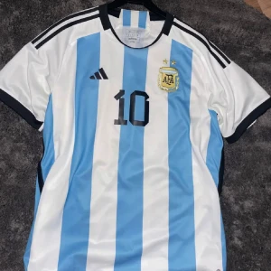 Argentina Messi fotbollströja  - Säljer en officiell Argentina fotbollströja från World Cup 2022 Adidas i storlek XL men passar även L, slim fit. Tröjan har klassiska blå och vita vertikala ränder, svarta detaljer och nummer 10 med Messi på ryggen. Materialet är lätt och andas, perfekt för match eller träning.