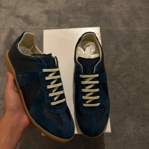Maison Margiela Gat Navy Blue - Säljer ett par Maison Margiela sneakers i mörkblå mocka och läder med beige snörning och gummisula. Klassisk siluett med rund tå och platt sula. Kommer med originalbox och dustbags. Perfekta för dig som gillar stilrena och lyxiga sneakers.
