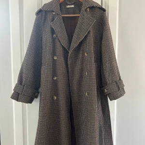 Rutig trenchcoat från Gina Tricot - Säljer en snygg rutig trenchcoat från Gina Tricot i bruna och beige toner. Jackan har dubbelknäppning, bälte i midjan och klassisk krage. Långa ärmar med knappar vid ärmslut. Perfekt för dig som vill sticka ut i höst och vår!