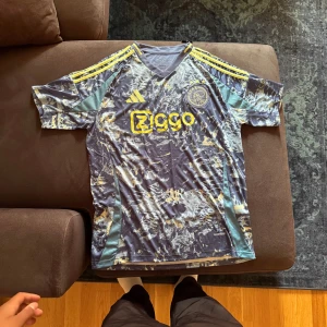 Ajax blå mönstrad fotbollströja Adidas - Ajax fotbollströja från Adidas med coolt abstrakt mönster i blått, grått och gult. Ziggo-logga och Ajax-emblem på bröstet, samt gula Adidas-ränder på axlarna. Tröjan har V-ringning och är tillverkad i lätt, ventilerande polyester.