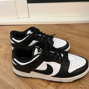 Nike Dunk Low svart/vit sneakers - Säljer ett par Nike Dunk Low sneakers i svart och vitt. Skorna har klassisk låg siluett, svart swoosh och snörning. Ovandelen är i skinn och de har en platt sula med rund tå. Perfekta för dig som gillar streetstyle och vill ha en ikonisk look.