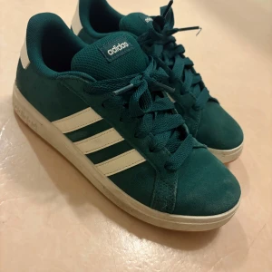 Adidas sneakers i mörkgrön mocka - Snygga Adidas sneakers i mörkgrön mocka med klassiska vita ränder på sidorna och vit sula. Skorna har snörning och rund tå, samt Adidas-logga på plösen och hälen. Perfekta för dig som gillar en sportig och stilren look.