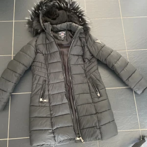 Svart lång dunjacka Polo Club  - Svart lång dunjacka från Polo Club med huva och fuskpäls, quiltad design och två dragkedjefickor. Jackan har en snygg siluett och är perfekt för kalla dagar. Stängs med dragkedja hela vägen och har en klassisk look.