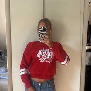 Röd croppad sweatshirt med tigertryck - Röd croppad sweatshirt  med vitt tigertryck framtill och  vita ränder på ärmarna. Tröjan har rund halsringning och långa ärmar, perfekt för en sportig och avslappnad stil.