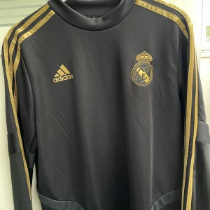 Svart Real Madrid långärmad tröja - Snygg svart långärmad fotbollströja från Adidas med Real Madrid-logga och guldiga detaljer. Tröjan har klassiska guldiga ränder längs ärmarna och Adidas-logga på bröstet. Tillverkad i mjukt och funktionellt material som passar perfekt för träning eller match.