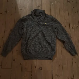Grå half zip tröja från Lyle & Scott - Mörkgrå tröja från Lyle & Scott med half zip och klassisk gul logga på bröstet. Tröjan har lång ärm, ribbade muddar och är tillverkad i ett mjukt stickat material. Perfekt för dig som gillar stilren och bekväm look.