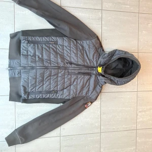 Cardigan från Parajumpers storlek L - Snygg svart cardigan från Parajumpers med huva och dragkedja framtill. Köpt i room 1006 i (Malmö emporia)Jackan har quiltad framsida, släta ärmar och två dragkedjefickor. Materialmix av tyg och puffer ger både värme och stil. Perfekt för dig som vill ha en snygg och stilren jacka. Ny pris för denna jacka ca 4300