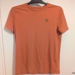 Orange t-shirt från Lyle & Scott 158/164 - Snygg orange t-shirt från Lyle & Scott med klassisk rund hals och korta ärmar. T-shirten har en gul broderad logga på bröstet och är gjord i mjuk bomull. Motsvarar strl 158/164