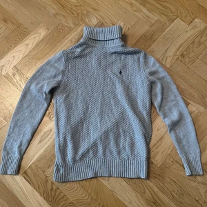 Grå polotröja från Morris - Snygg grå polotröja från Morris med stickad struktur och broderad svart lilja på bröstet. Tröjan har ribbade muddar och hög polokrage, perfekt för lager-på-lager. Passar dig som gillar klassisk men modern stil.