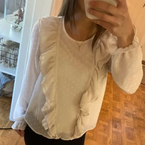 Vit blus med volangdetaljer - Supersöt vit blus med långa ärmar och volanger framtill. Blusen har diskret prickigt strukturmönster och är tillverkad i ett lätt, luftigt tyg. Perfekt för dig som gillar romantiska och feminina detaljer.