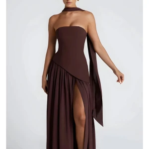 Brun långklänning med slits - Snygg brun axelbandslös långklänning med asymmetrisk midja och hög slits framtill. Klänningen har en elegant scarf-detalj runt halsen och ett flowy, plisserat kjolparti. Perfekt för dig som vill sticka ut med en stilren och modern look.