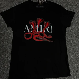 Svart Amiri t-shirt med röd ormprint - Svart t-shirt från Amiri med ett stort tryck av röda ormar och vit logga på bröstet. Klassisk passform och rund halsringning. Perfekt för dig som vill sticka ut med en edgy vibe.