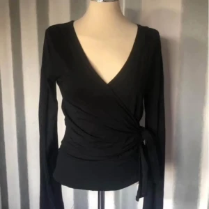 Svart omlottopp med knytband - Svart omlottopp med lång ärm och djup v-ringning. Toppen har knytband i sidan som ger en snygg passform och trendig look. Perfekt att styla med jeans eller kjol för en cool outfit. Storlek 36/38 , viscose mjuk bomulls känsla . Nyskick 