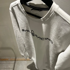 Vit Sail Racing sweatshirt i bomull - Vit sweatshirt från Sail Racing med svart logotyptryck över bröstet. Tröjan har rund halsringning, långa ärmar och är tillverkad i mjuk bomull. Enkel och clean design som passar perfekt till jeans eller joggers.