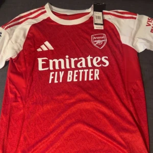 Arsenal Gyökeres 14 matchtröja Adidas - Snygg Arsenal fotbollströja med Gyökeres och nummer 14 på ryggen. Röd kropp med vita ärmar och detaljer, Adidas-logga och Emirates Fly Better tryck på bröstet. Tröjan har ett subtilt mönster över hela framsidan och är tillverkad i lätt, ventilerande material.