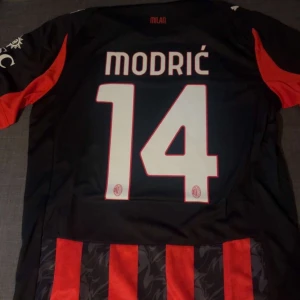 AC Milan Modric 14 Puma fotbollströja - Snygg AC Milan fotbollströja med Modric och nummer 14 på ryggen. Tröjan är svart och röd med klassiska vertikala ränder, Puma-logga och Emirates Fly Better tryck på bröstet. Materialet är lätt och andas, perfekt för match eller träning.