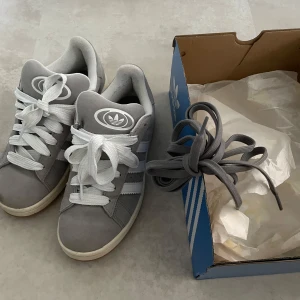 Adidas Campus 00s grå sneakers - Säljer ett par Adidas Campus 00s sneakers i grå mocka med vita detaljer och klassiska tre ränder på sidan. Skorna har vit sula, rund tå och snörning. Medföljer extra grå skosnören. Perfekta för dig som gillar retrovibbar och streetstyle.