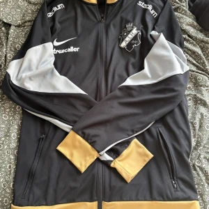 AIK Nike ziptröja svart/gul XL - AIK ziptröja från Nike i svart med gula muddar och detaljer samt gråa partier på ärmarna. Broderad AIK-logga på bröstet, dragkedja hela vägen och två fickor med dragkedja. Tillverkad i Dri-FIT polyester, perfekt för fotboll eller träning.