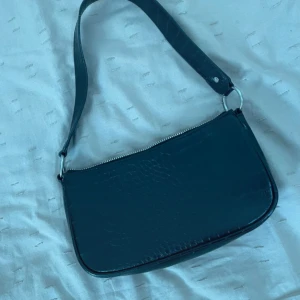 Svart axelväska i croco-imitation - Snygg svart axelväska med croco-mönstrad yta och silvrig dragkedja. Väskan har en bred axelrem och är tillverkad i syntetmaterial som ger en lyxig känsla. Perfekt storlek för det viktigaste och en stilren look.