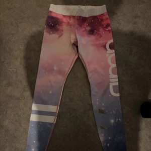 Galaxy leggings från aim'n, S - Snygga leggings från aim'n med galaktiskt mönster i rosa, lila och blå toner. De har bred vit midja, vita ränder på benen och stor logga på sidan. Materialet är stretchigt och består av 73% polyester och 27% spandex – perfekta för träning eller chill., som man ser är de en fläck där i fram.