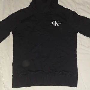 Svart hoodie från Calvin Klein Jeans - Svart hoodie från Calvin Klein Jeans med vit logga på bröstet. Klassisk modell med huva och ribbade muddar. Perfekt för en chill och stilren look. Materialet känns mjukt och bekvämt mot huden. Passar dig som är runt 150 cm!