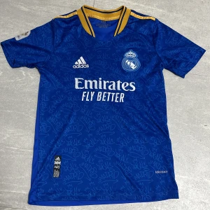 Real Madrid Benzema blå matchtröja - Snygg blå Real Madrid fotbollströja från Adidas med Benzema och nummer 9 på ryggen. Tröjan har gula detaljer vid krage och axlar, samt grafiskt mönster med Madrid-tryck över hela. Kortärmad och tillverkad i lätt funktionsmaterial (Aeroready). Perfekt för dig som älskar fotboll och Real Madrid.