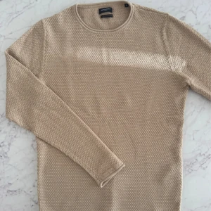 Beige stickad tröja Jack & Jones - Snygg beige stickad tröja från Jack & Jones med rund halsringning och lång ärm. Tröjan har en grovstickad struktur som ger en cool och avslappnad vibe. Perfekt att slänga på sig när det är lite kyligare ute. Perfekt skick. 