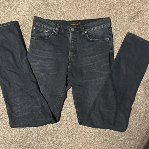 Mörkblå jeans från Nudie Jeans Co - Snygga mörkblå jeans från Nudie Jeans Co med klassisk femficksdesign och diskreta slitningar. Jeansen har raka ben och broderade bakfickor med Nudie-loggan. Perfekta för dig som gillar stilrena och tidlösa jeans.