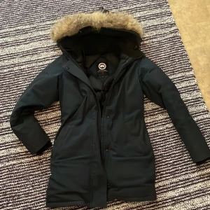 Marinblå vinterjacka med päls från Canada Goose - Säljer en marinblå Canada Goose vinterjacka  med äkta päls på huvan. Jackan har dragkedja framtill och flera praktiska fickor. Insidan är quiltad och jackan är perfekt för kalla vinterdagar. Snygg och tidlös modell som håller dig varm hela säsongen, den är använd 2 vintrar jackan är hel och funkar jättebra bara att på slutet av ärmarna å Ede lite slitet inne i ärmarna och vid snöretna vid luvan e lite slitet oxå det är inget märkvärdigt jag säljer den då den har blivit för liten för mig! :)