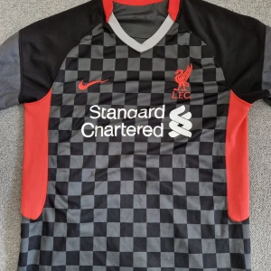 Liverpool rutiga bortatröja Nike - Liverpool FC bortatröja från Nike med svart och grått rutmönster, röda detaljer och klubbmärke broderat på bröstet. Kortärmad modell med V-ringning och tryckt sponsorlogga på magen. Perfekt för dig som vill sticka ut på läktaren eller i stan.