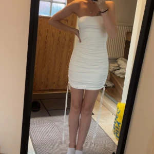 Vit axelbandslös kortklänning - Säljer en vit, tight och axelbandslös kortklänning med snygg rynkdetalj och knytband på sidorna. Klänningen har en figurnära passform och är perfekt för dig som vill ha en stilren och trendig look. Materialet är stretchigt. Aldrig använd då jag råka få hem 2 st likadana. 