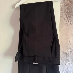 Svarta kostymbyxor från Tiger of Sweden - Stilrena svarta kostymbyxor från Tiger of Sweden i modellen Noowa  med raka, vida ben och klassisk passform. Byxorna har hög midja och är tillverkade i ett mjukt tyg som ger en dressad look. Perfekta för dig som gillar en clean och tidlös stil.