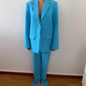 Turkos kostym med kavaj och byxa - Snygg turkos kostym med klassisk kavaj och matchande raka kostymbyxor. Kavajen har två knappar framtill, breda slag och fickor med lock. Byxorna är höga i midjan och har en stilren, rak passform. Perfekt för dig som vill sticka ut med färg.