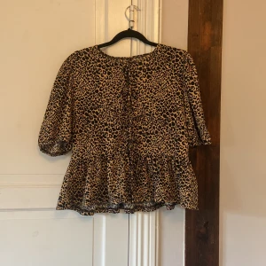 Leopardmönstrad blus med volang - Blus med leopardmönster i beige och svart. Modellen har puffiga armar och en volang.