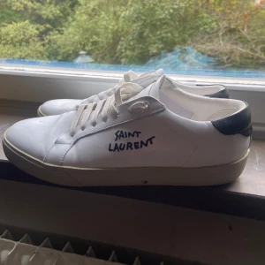 Saint Laurent skor - Snygga vita sneakers från Saint Laurent med svart detalj på hälen och broderad logga på sidan. Skorna är i skinn och har klassisk snörning samt rund tå. Skorna är använda i ca 1 månad skick 8/10