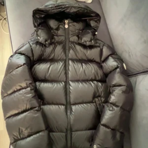 Svart Moncler pufferjacka med huva - Säljer en svart pufferjacka från Moncler med glansig finish och stor huva. Jackan har dragkedja framtill och klassisk Moncler-logga på ärmen. Perfekt för kalla dagar och riktigt snygg streetstil.