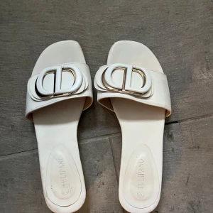 Vita sandaler med metallspänne från Tulipano - Säljer ett par stilrena vita sandaler från Tulipano med bred rem och stort dekorativt metallspänne framtill. Platt sula och öppen tå gör dem perfekta för varma dagar. Enkla att matcha till olika outfits.