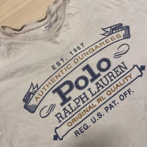 Ralph Lauren t shirt - Beige/ lite vit t-shirt från Polo Ralph Lauren med stort tryck i blått och brunt framtill. Klassisk rund halsringning och korta ärmar. Perfekt för en avslappnad och stilren look nu till sommaren.                 Storlek (m) men passar som (s).