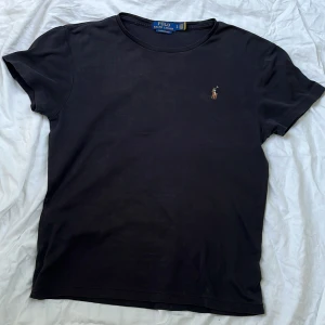 Svart t-shirt från Polo Ralph Lauren - Passar 13-14 åringar. Kostar 1200kr ny. Svart t-shirt från Polo Ralph Lauren med klassisk broderad logga på bröstet. Modellen är custom slim fit och har korta ärmar. Perfekt för en stilren och enkel look.
