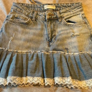 Jeanskjol med volang och spetskant - Söt jeanskjol med volang och vit spetskant längst ner. Kjolen har slitna detaljer, fickor fram och bak samt en extra mörkblå bakficka. Perfekt för dig som gillar en lekfull och trendig stil.