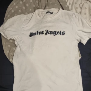 Vit t-shirt från Palm Angels - Säljer en vit t-shirt från Palm Angels med svart logotyptext på bröstet. Klassisk rund halsringning och korta ärmar. Perfekt för dig som gillar streetwear och vill ha en stilren look. Krympt i stl till L-XL