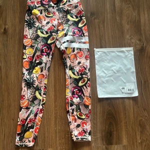 Färgglada leggings med fruktmönster från Stronger - Säljer ett par färgglada leggings från Stronger med tropiskt fruktmönster i rosa, gult, grönt och orange. Byxorna har vita ränder på låret och är perfekta för dig som vill ha en färglad outfit. Superstretchigt material och hög midja! 💓