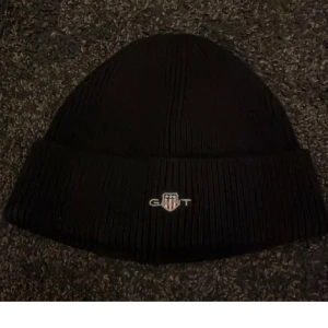 Svart ribbad mössa från GANT - Svart ribbstickad mössa från GANT med broderad logotyp framtill. Klassisk modell med uppvikt kant, perfekt för kyliga dagar.