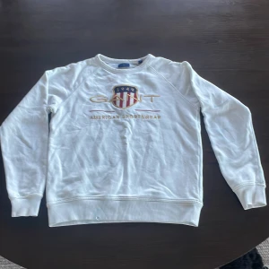 Vit sweatshirt från GANT - Vit sweatshirt från GANT med broderad logga och texten 'American Sportswear' framtill. Tröjan har långa ärmar, rund halsringning och ribbade muddar vid ärmslut och nederkant. Klassisk och stilren design med broderad detalj. DEN HAR BLÅA FLÄCKAR HÄR OCH DÄR!!!