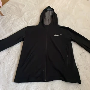Svart hoodiejacka från Nike Dri-Fit - Svart hoodiejacka från Nike med dragkedja och Dri-Fit-material. Jackan har lång ärm, huva och en diskret Nike-logga på bröstet. Perfekt för träning eller chill dagar. Snygg och enkel design.